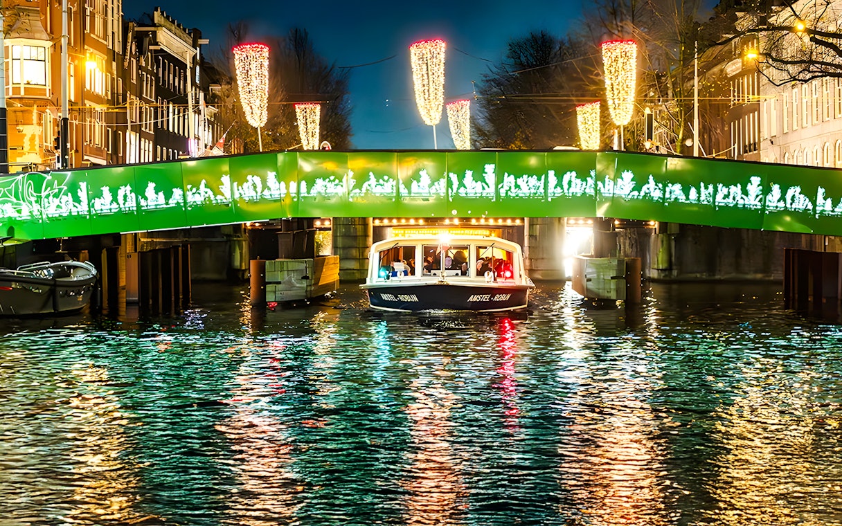 Boat passing under illuminated bridge during Amsterdam Light Festival evening cruise.