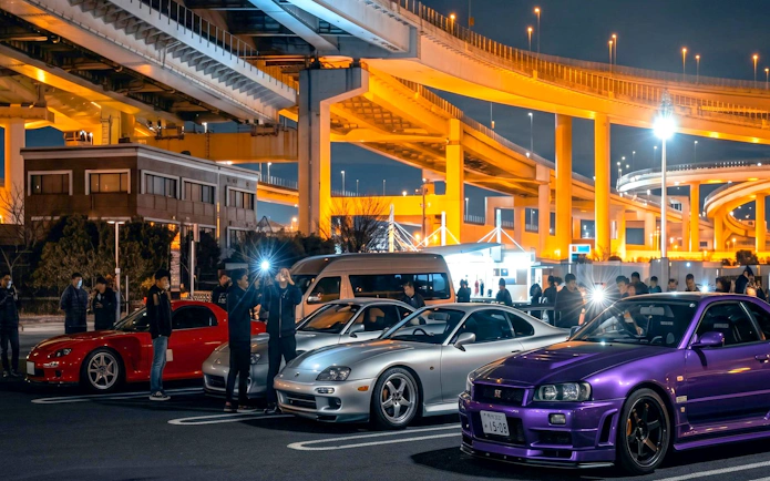 Skyline R34 and other cars at Daikoku PA car meeting, Japan, under illuminated highway.