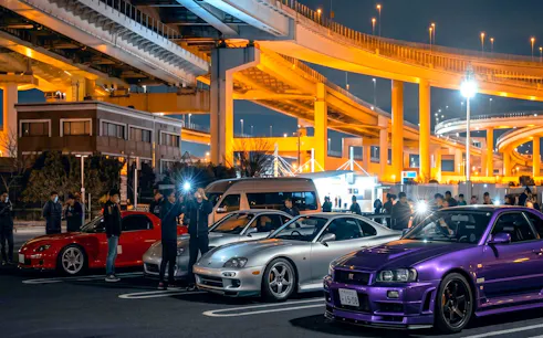 Skyline R34 and other cars at Daikoku PA car meeting, Japan, under illuminated highway.