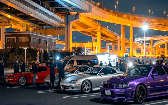 Skyline R34 and other cars at Daikoku PA car meeting, Japan, under illuminated highway.
