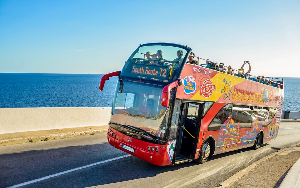 Open-top tour bus on coastal road in Malta with passengers enjoying sea views.