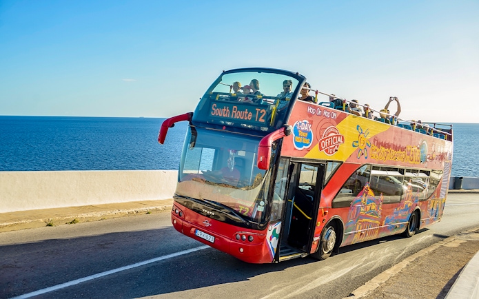 Open-top tour bus on coastal road in Malta with passengers enjoying sea views.