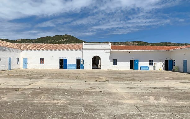 Courtyard of historic building in Asinara National Park, Sardinia, with blue doors and mountain backdrop.