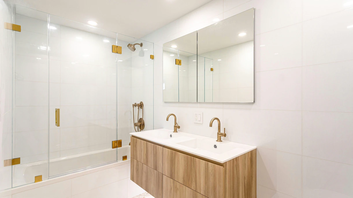 Modern white bathroom with double vanity, glass shower, and brass fixtures.