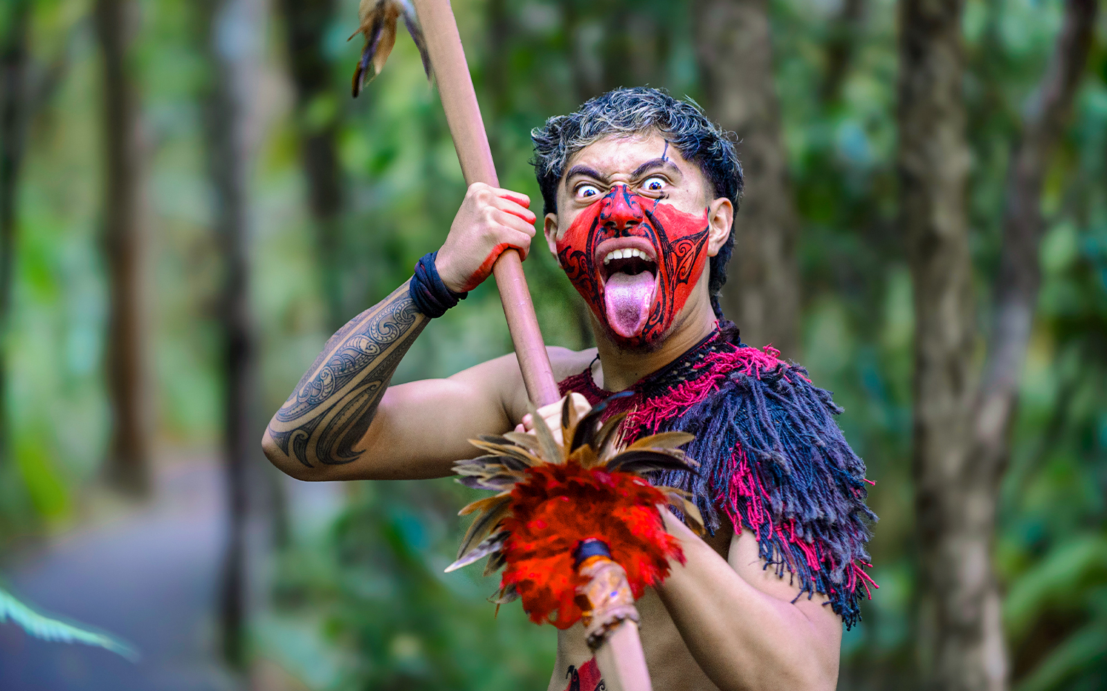 Warrior performing traditional challenge with face paint and spear in forest setting.