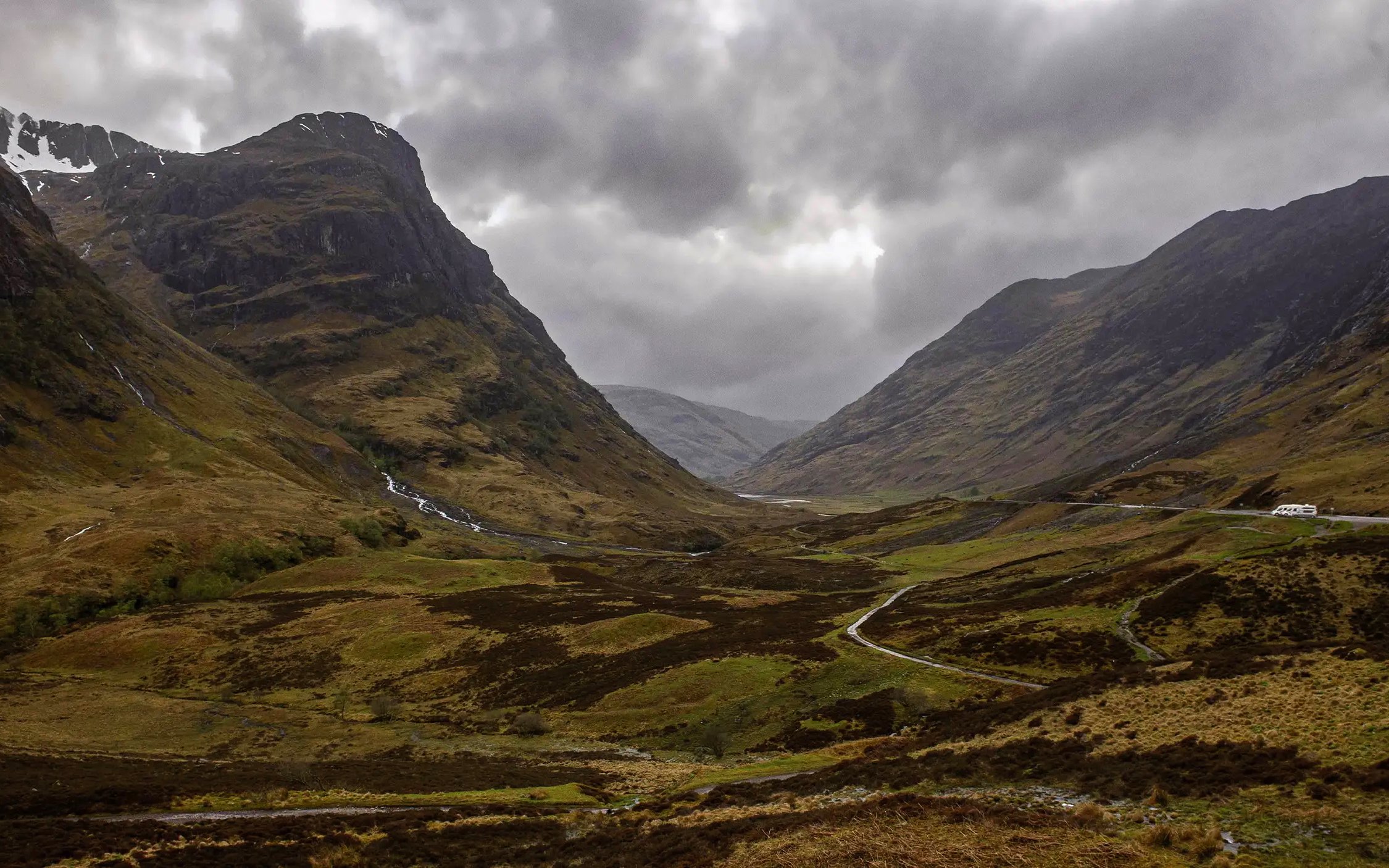 Mountainous landscape of Glencoe, Scotland, with winding road and cloudy sky.