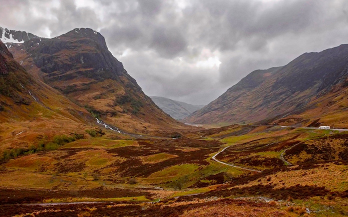 Mountainous landscape of Glencoe, Scotland, with winding road and cloudy sky.