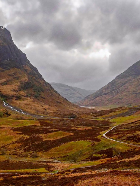 Mountainous landscape of Glencoe, Scotland, with winding road and cloudy sky.