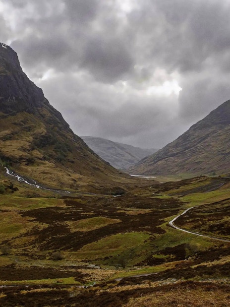 Mountainous landscape of Glencoe, Scotland, with winding road and cloudy sky.