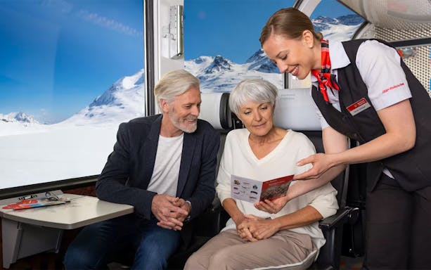 Passengers on Bernina Express receiving brochure from train staff with snowy Alps view.