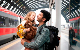 Father holding child with teddy bear at a train station platform.