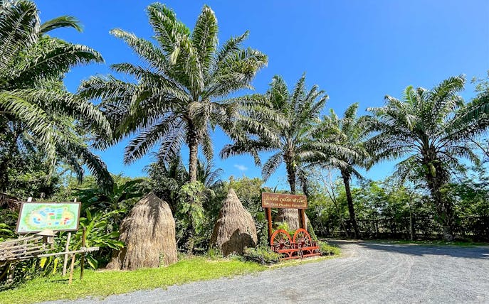 Thai farm entrance with palm trees and traditional haystacks, part of a guided cooking class tour.