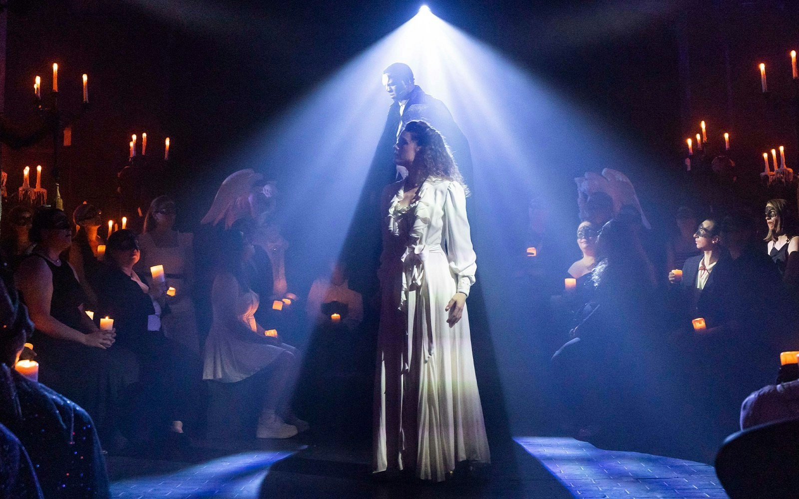 Masquerade Broadway stage performance with actors in spotlight and audience holding candles.