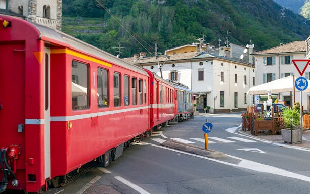 Bernina train passing through Tirano city street, Italy, with buildings and mountains in the background.