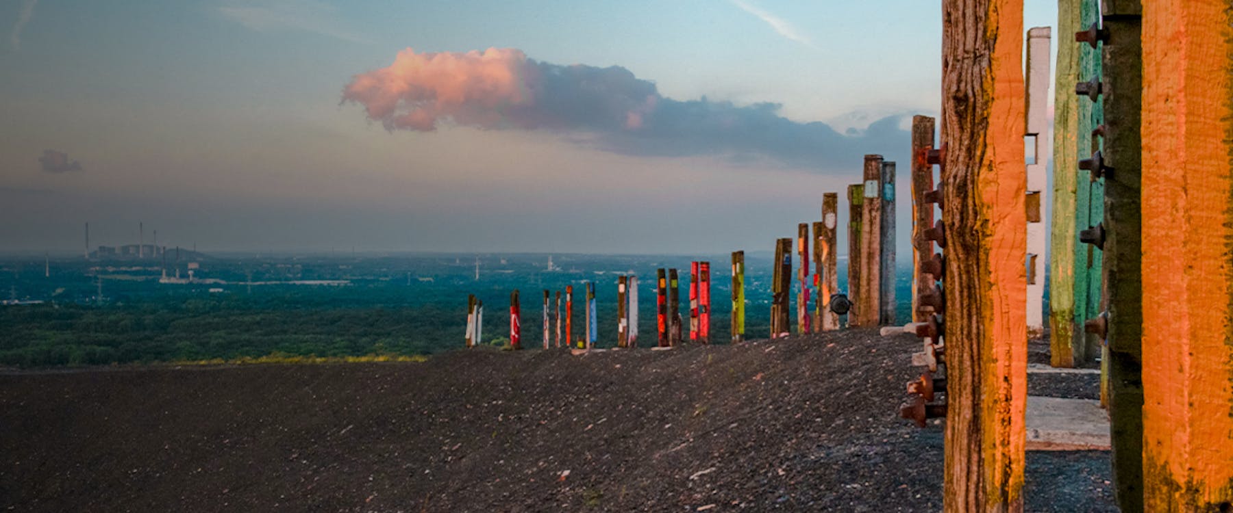 Colorful wooden poles on a hilltop in Bottrop, Germany, overlooking a vast landscape.