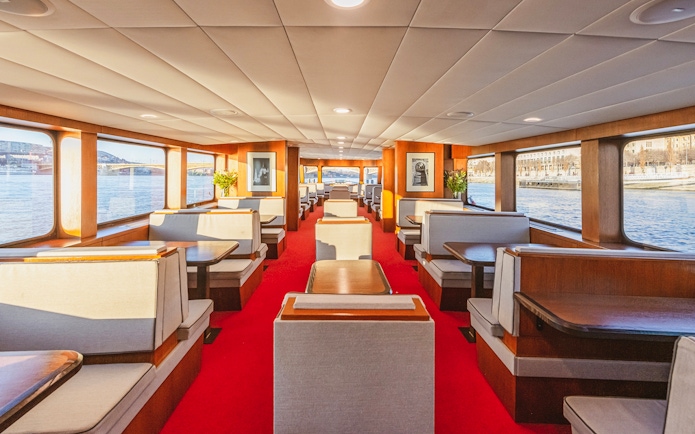 Interior of a sightseeing cruise with large windows and dining booths along a river.