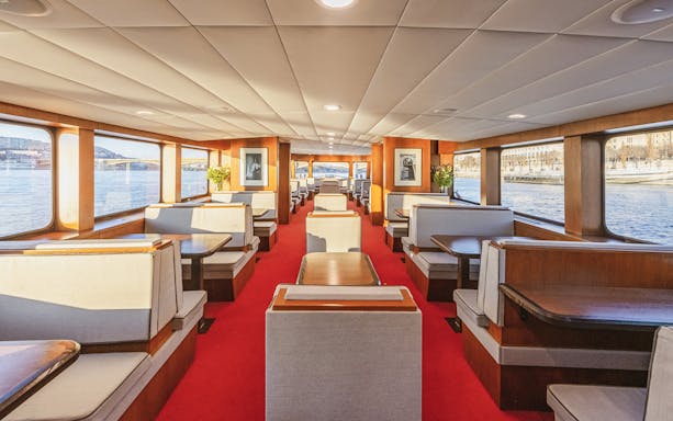 Interior of a sightseeing cruise with large windows and dining booths along a river.