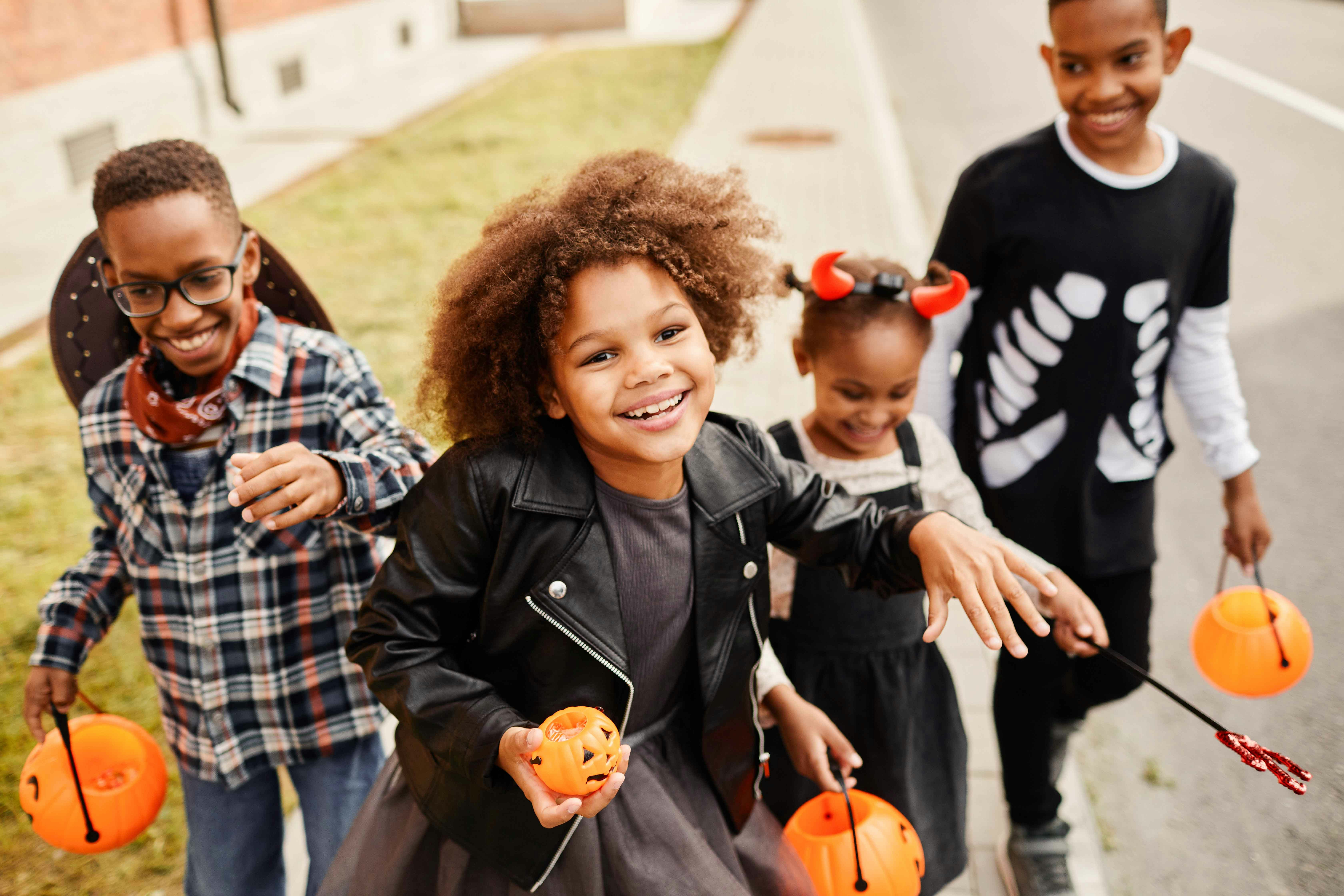 Children in Halloween costumes holding pumpkin buckets on a sidewalk.