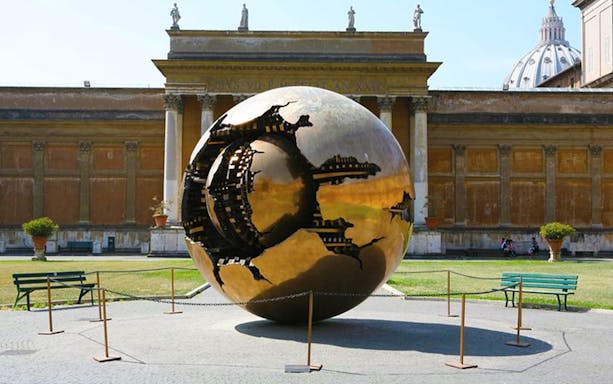 Sphere Within Sphere sculpture at Vatican Museums, Rome, with St. Peter's Basilica dome in background.