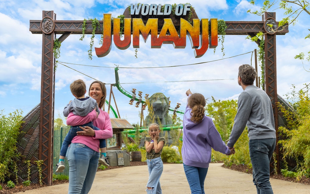 Family entering World of Jumanji theme park under entrance sign with roller coaster in background.