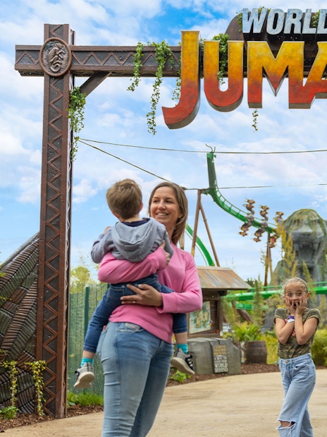Family entering World of Jumanji theme park under entrance sign with roller coaster in background.