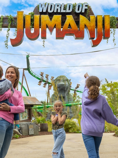 Family entering World of Jumanji theme park under entrance sign with roller coaster in background.