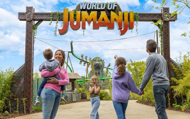 Family entering World of Jumanji theme park under entrance sign with roller coaster in background.