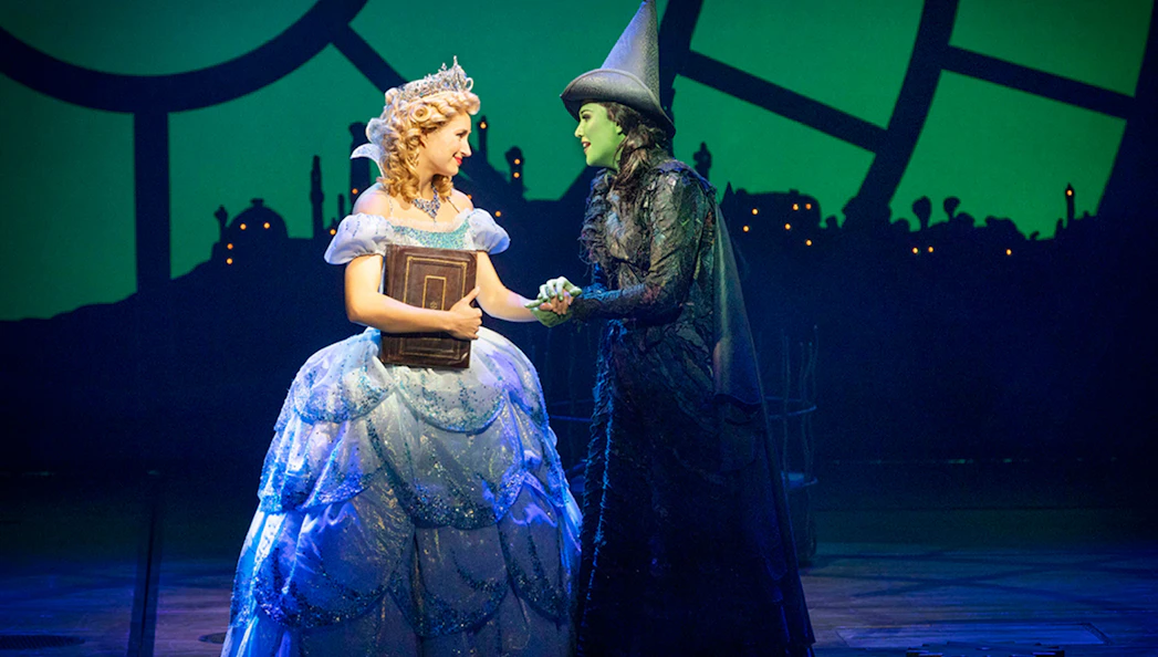 Two characters from Wicked musical on stage, one in a blue gown, the other in a black hat.