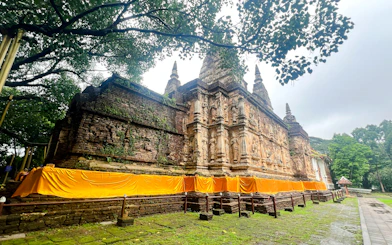 Ancient temple with intricate carvings and orange drapery in Chiang Mai, surrounded by lush greenery.