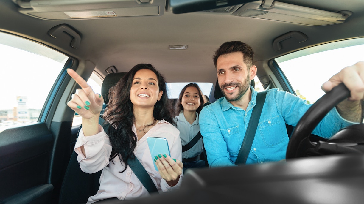 Family in car driving to travel destination, smiling and pointing ahead.