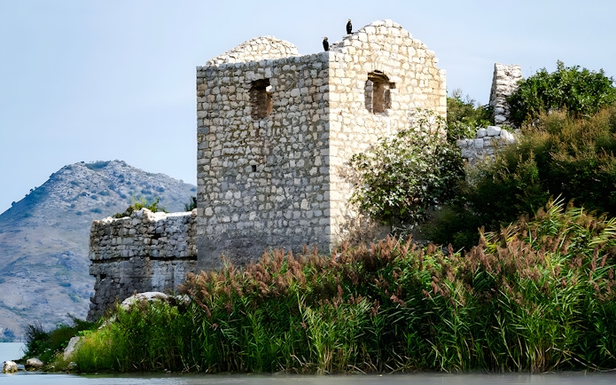 Grmožur Fortress ruins by a lake with mountain backdrop.