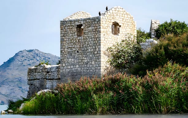 Grmožur Fortress ruins by a lake with mountain backdrop.