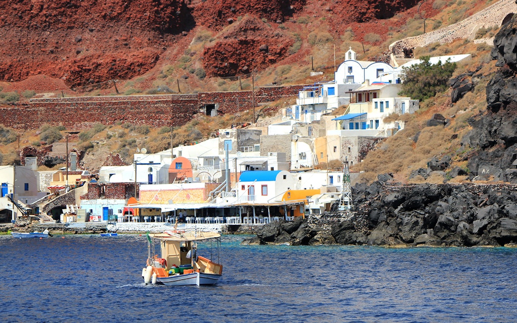 Fishing boat near colorful houses on Thirassia Island, Santorini, with volcanic cliffs in the background.