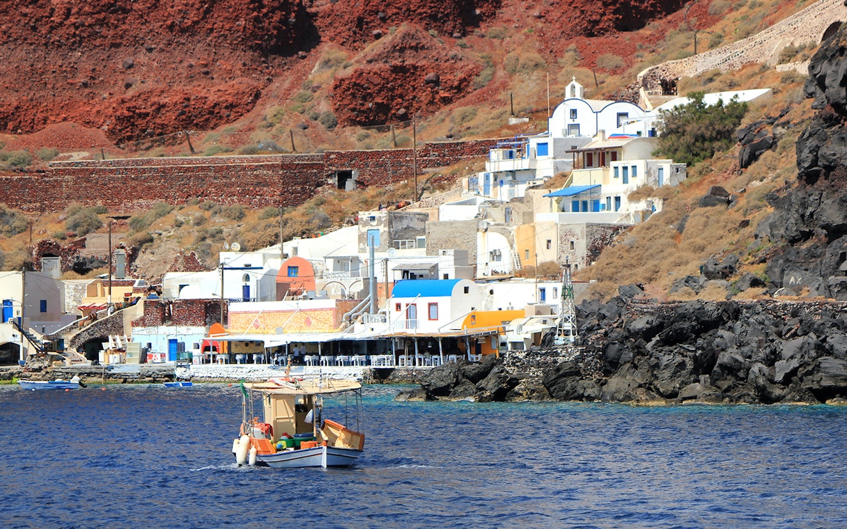 Fishing boat near colorful houses on Thirassia Island, Santorini, with volcanic cliffs in the background.