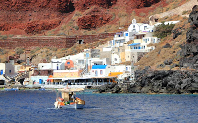 Fishing boat near colorful houses on Thirassia Island, Santorini, with volcanic cliffs in the background.