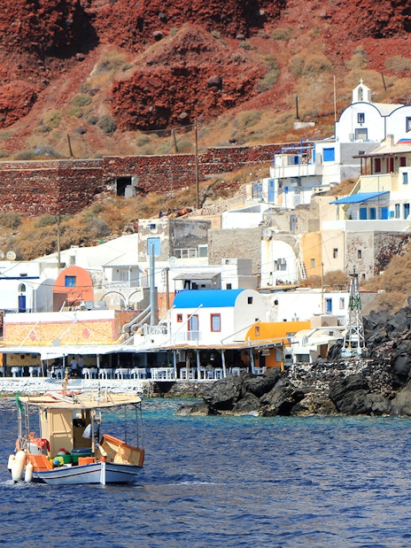Fishing boat near colorful houses on Thirassia Island, Santorini, with volcanic cliffs in the background.