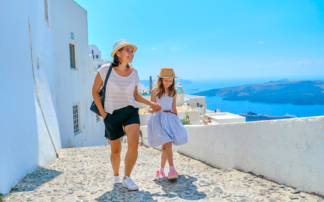 Mother and daughter walking on a cobblestone path in Santorini, Greece, with sea view.