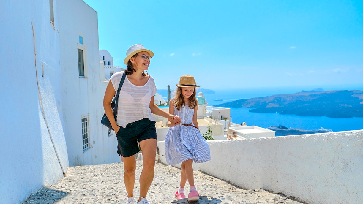Mother and daughter walking on a cobblestone path in Santorini, Greece, with sea view.