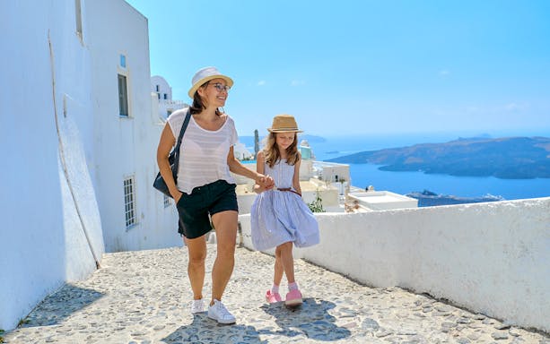 Mother and daughter walking on a cobblestone path in Santorini, Greece, with sea view.