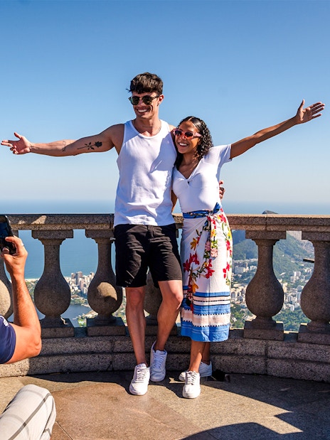 Couple posing at Christ the Redeemer viewpoint, Rio de Janeiro, with cityscape in background.