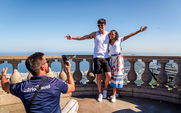 Couple posing at Christ the Redeemer viewpoint, Rio de Janeiro, with cityscape in background.
