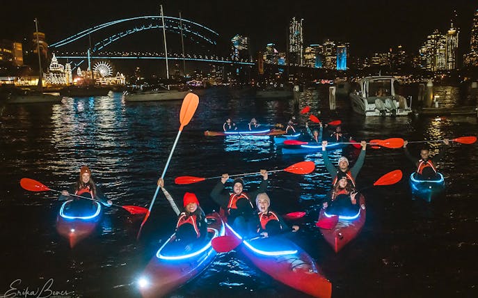 Kayakers paddle near Sydney Harbour Bridge lit up for Vivid festival at night.
