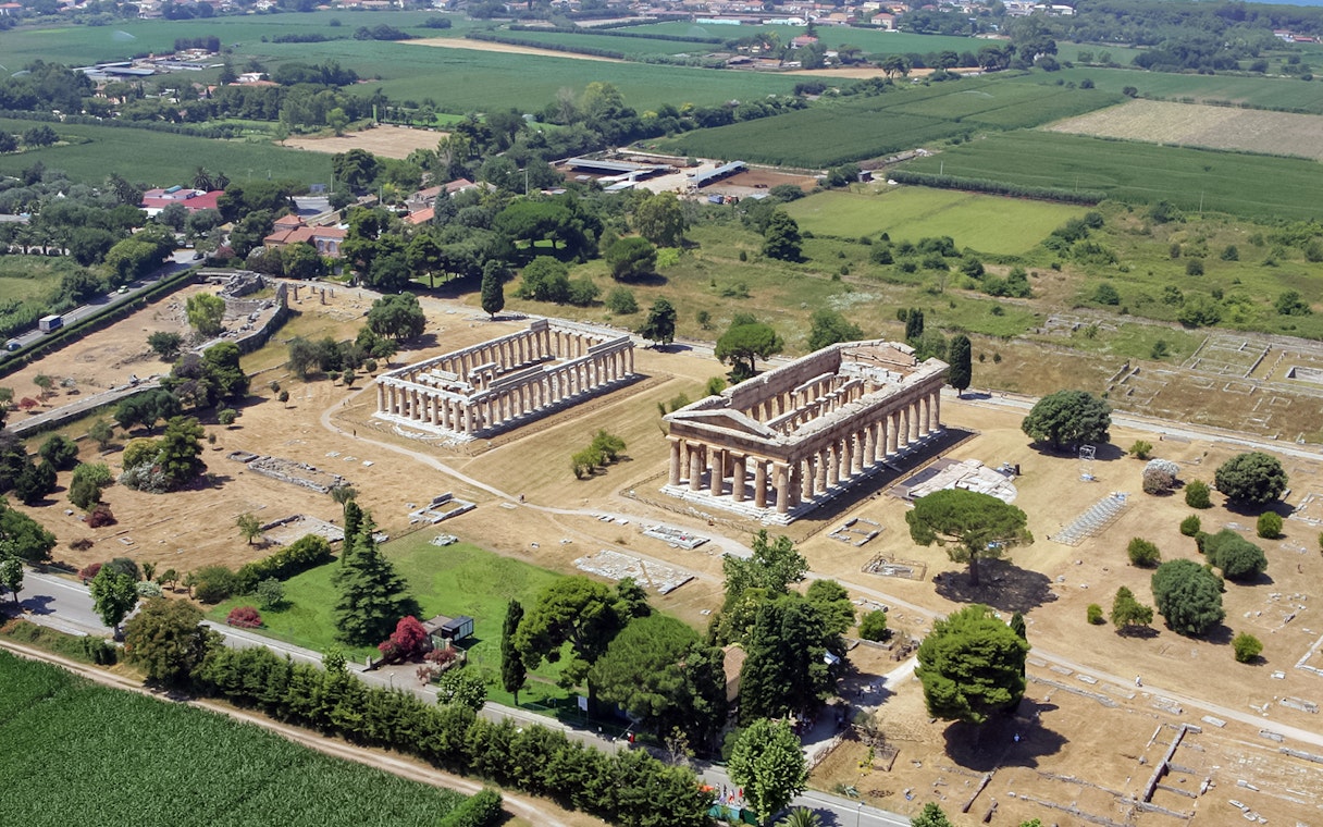 Aerial view of ancient Greek temples in Paestum, Italy, surrounded by green fields.