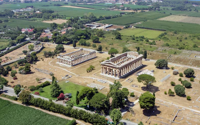 Aerial view of ancient Greek temples in Paestum, Italy, surrounded by green fields.