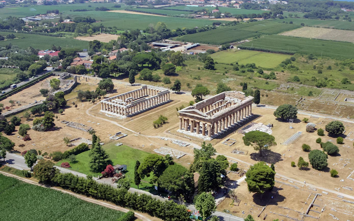 Aerial view of ancient Greek temples in Paestum, Italy, surrounded by green fields.