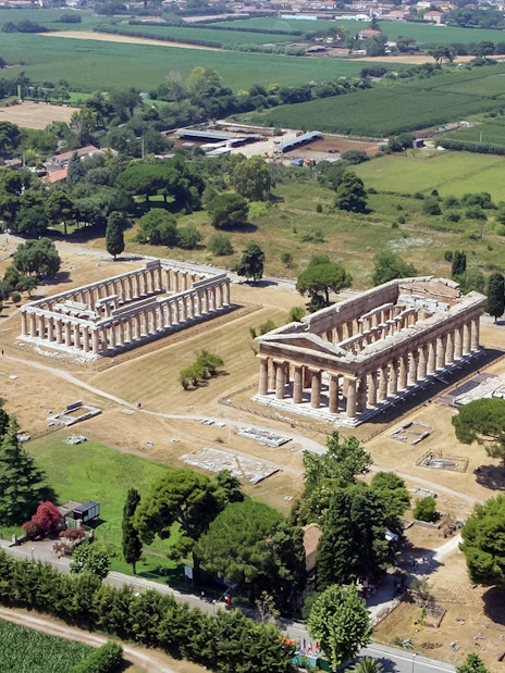 Aerial view of ancient Greek temples in Paestum, Italy, surrounded by green fields.