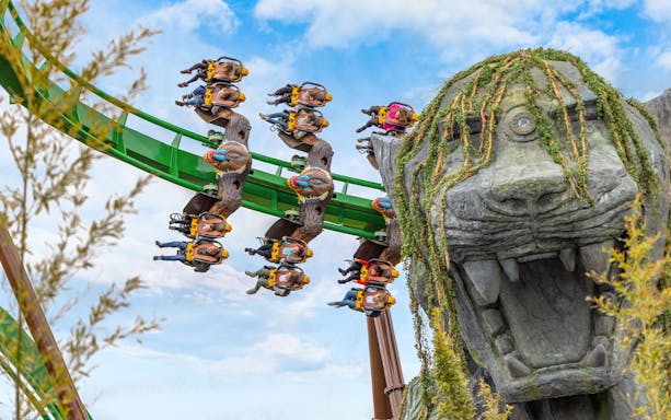 Guests on a Jumanji-themed roller coaster in a jungle setting with a giant stone lion head.