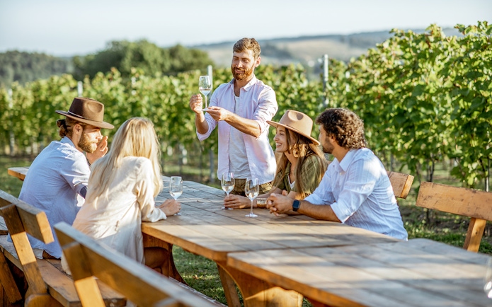 Young people enjoying wine and conversation at a vineyard table.