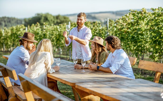 Young people enjoying wine and conversation at a vineyard table.