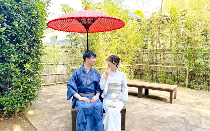 Couple in traditional Japanese kimonos sitting under a red umbrella in a garden.